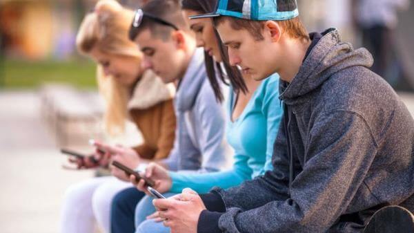 4 jongeren zitten buiten naast elkaar op een bank. Alle vier zitten ze in de houding die een text-neck kan veroorzaken.